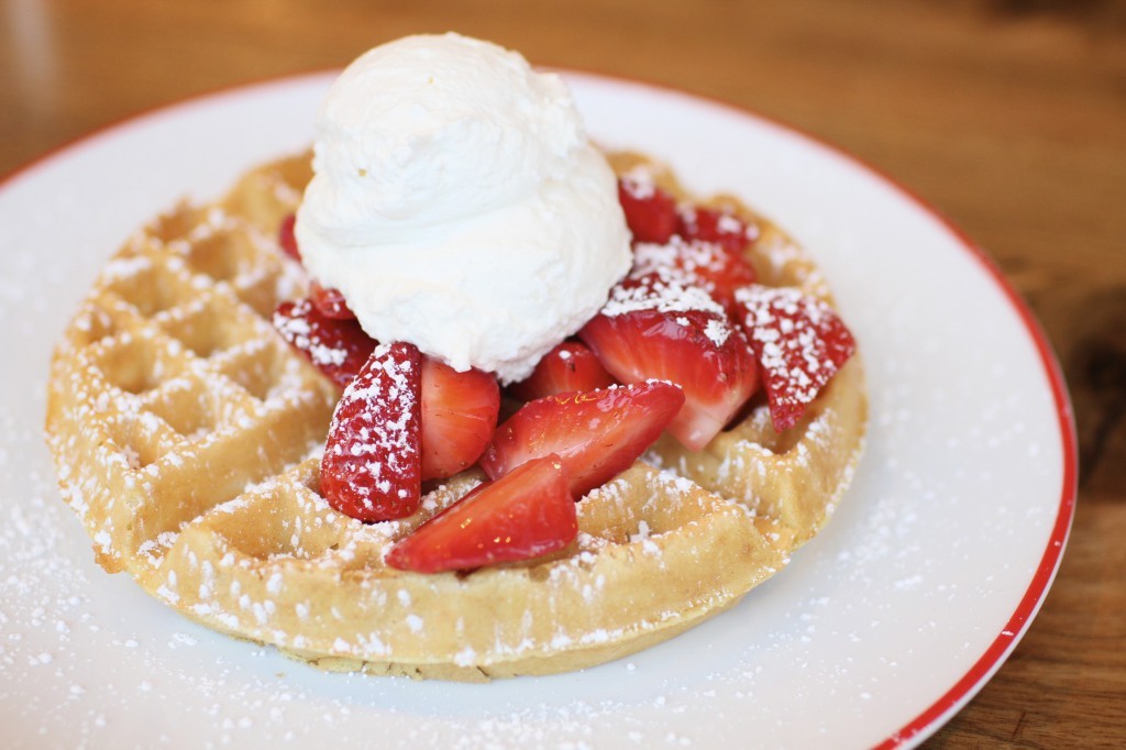 Farmhouse waffles topped with fresh strawberries and a dusting of powdered sugar, served on a white plate.