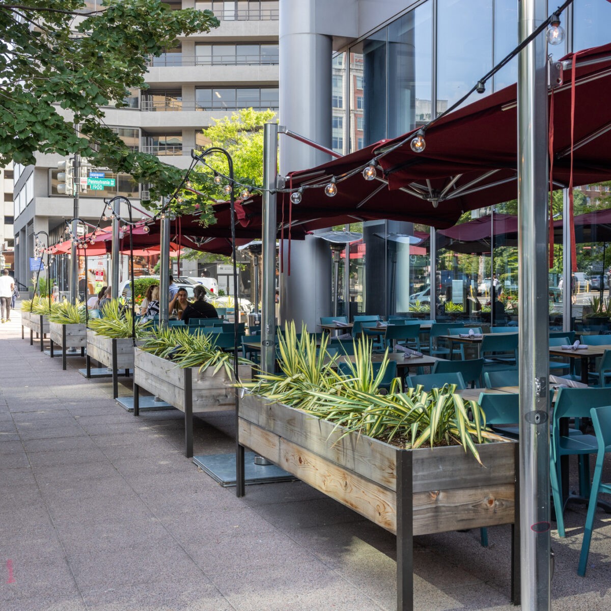 Street-side patio dining setup at Founding Farmers DC with teal chairs, red umbrellas, and leafy planters along the sidewalk.